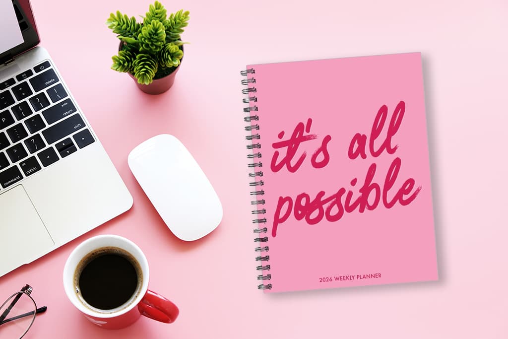 Pink 2026 weekly planner on a pink desk next to a laptop, coffee, and plant—symbolizing productive planning habits