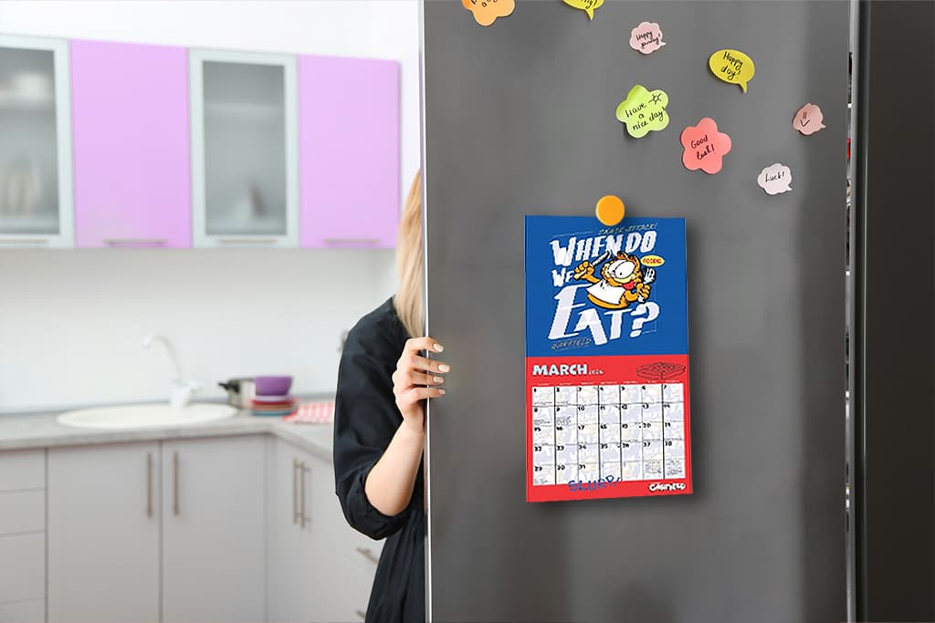 Mini wall calendar with cartoon design hanging on a fridge in a modern kitchen, surrounded by colorful sticky notes